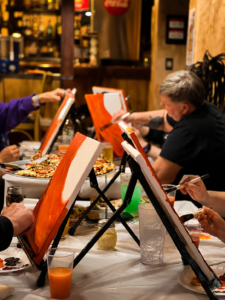 Adults painting orange fall canvases together at a cozy, beginner-friendly Artvana paint and sip event.”