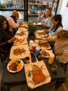 Adults painting fall pumpkin tote bags together at a beginner-friendly Artvana painting event with autumn colors and art supplies.”